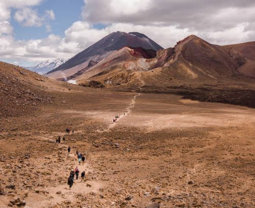 View from above to volcanic valley of Tongariro National Park in New Zealand, down there is a path with silhouettes of hikers, in a distance on the horizon, peaks of a crater